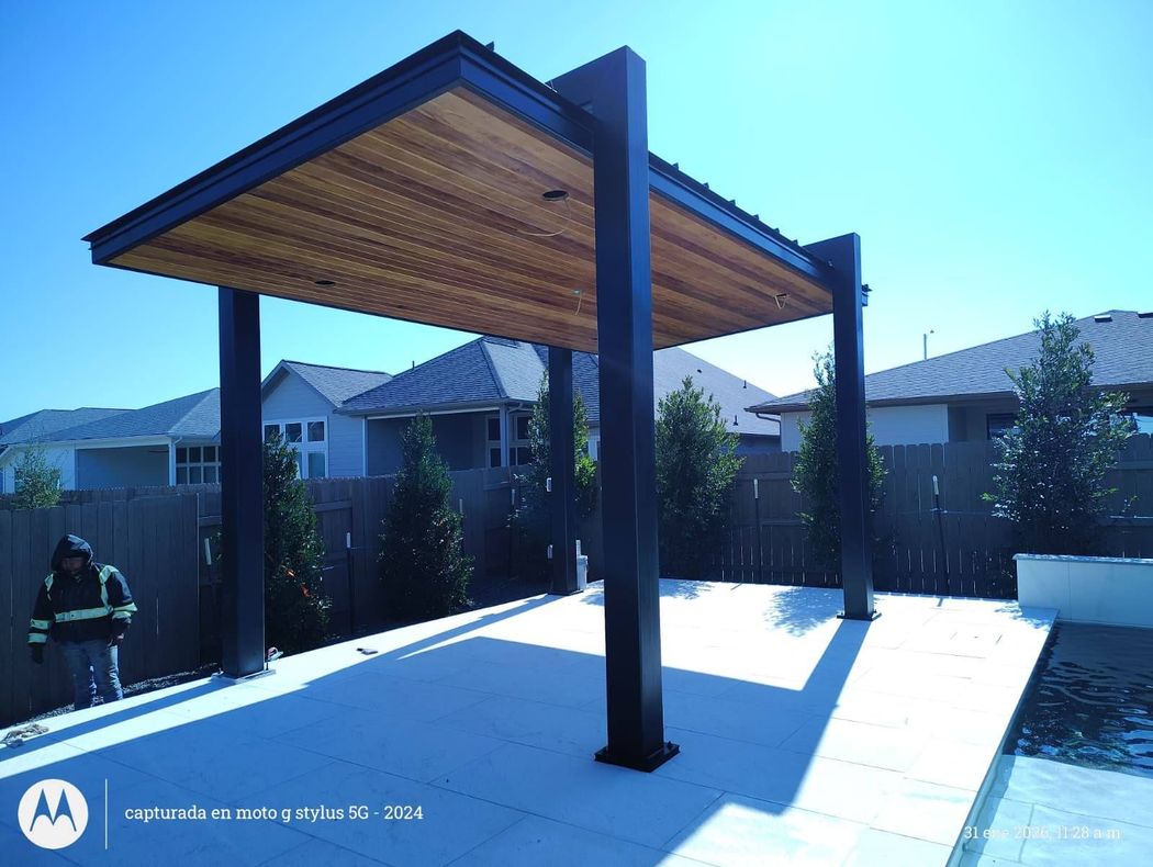 A modern black-framed patio pavilion with a wooden ceiling stands over a light-colored paved area in a backyard. A modern black-framed patio pavilion with a wooden ceiling stands over a light-colored paved area in a backyard.