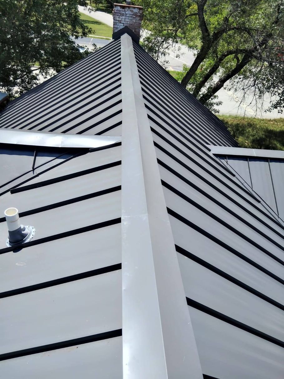 A top-down view of a dark metal roof ridge, showing the seam and surrounding panels leading to a brick chimney. A top-down view of a dark metal roof ridge, showing the seam and surrounding panels leading to a brick chimney.