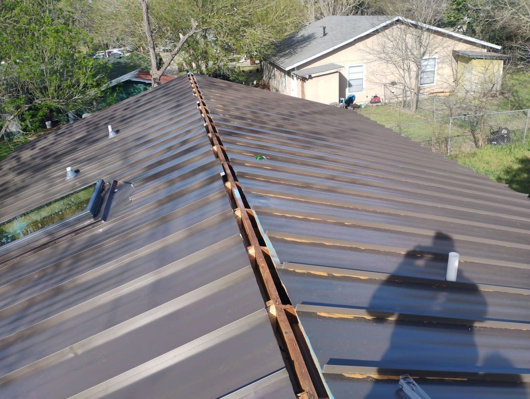 A high-angle view of a brown metal roof under repair, showing an open ridge vent strip and the shadow of the photographer. A high-angle view of a brown metal roof under repair, showing an open ridge vent strip and the shadow of the photographer.