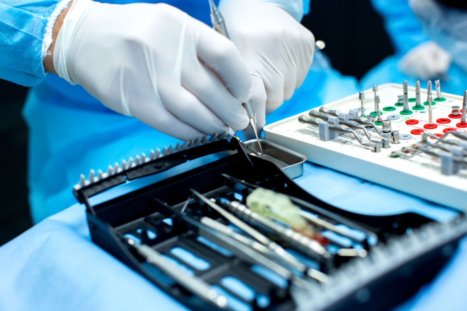 Surgeon's gloved hands holding a dental tool near an open surgical instrument tray with various tools.