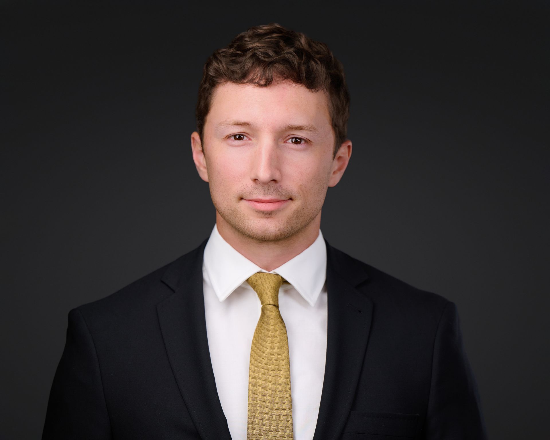 Man in suit and tie, smiling at the camera, against a dark gray background.