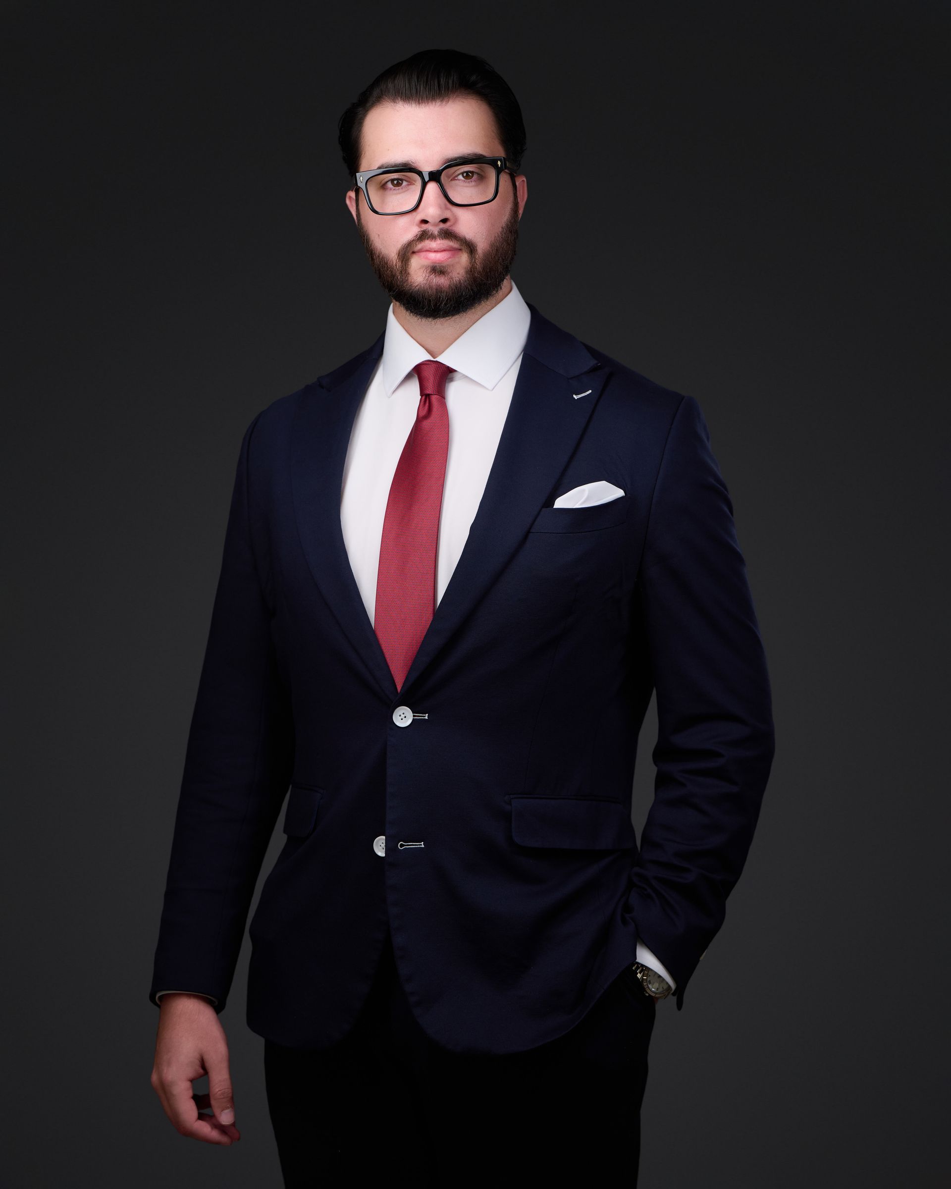 Man in navy suit, white shirt, red tie, and glasses poses against a dark backdrop.