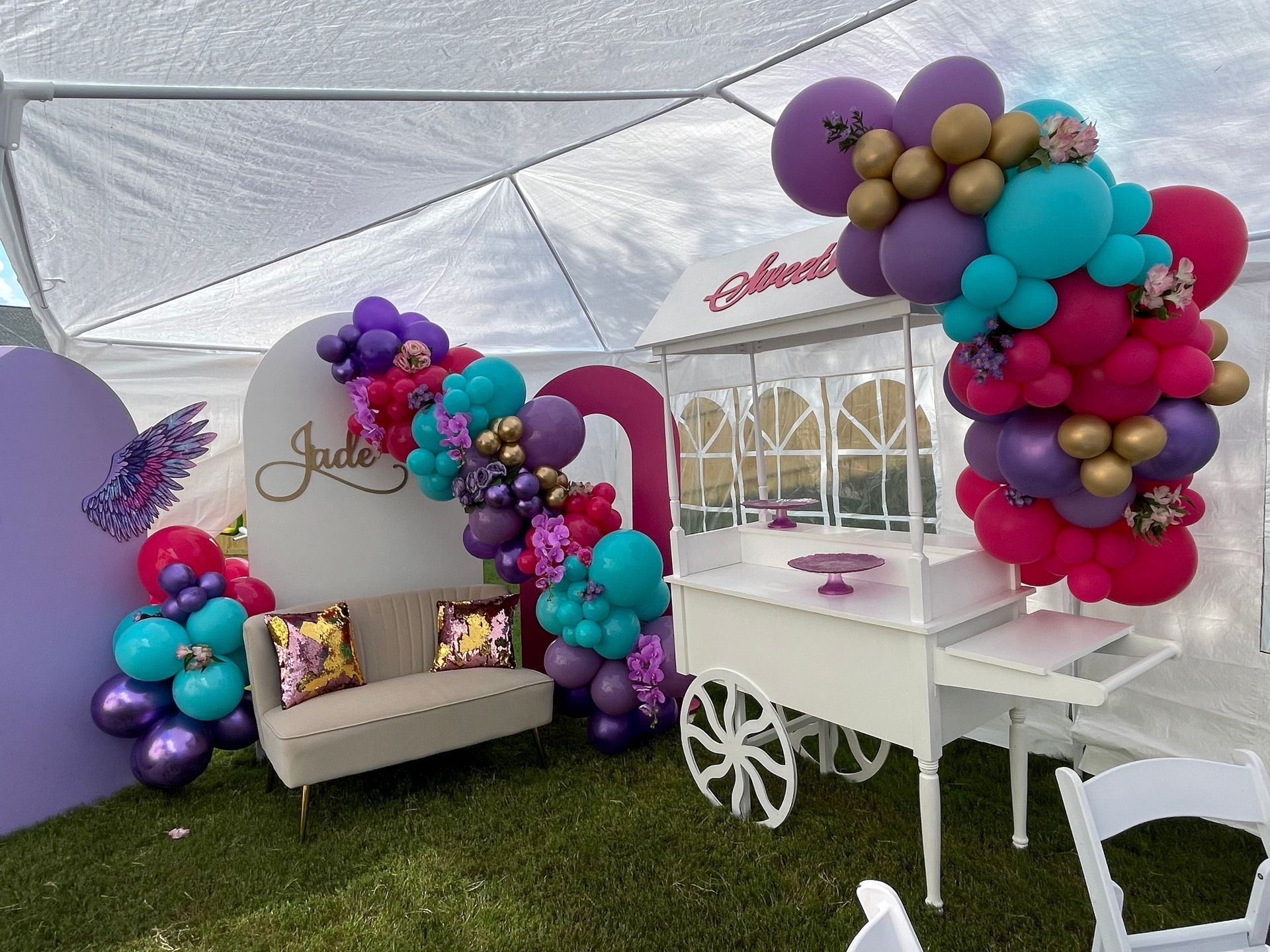 Colorful balloon garland around white treat cart and seating for whimsical outdoor kids party or festival