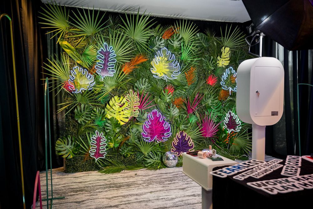 Tropical greenery photo booth backdrop with palm leaves, colorful floral accents, and selfie station setup for party rentals