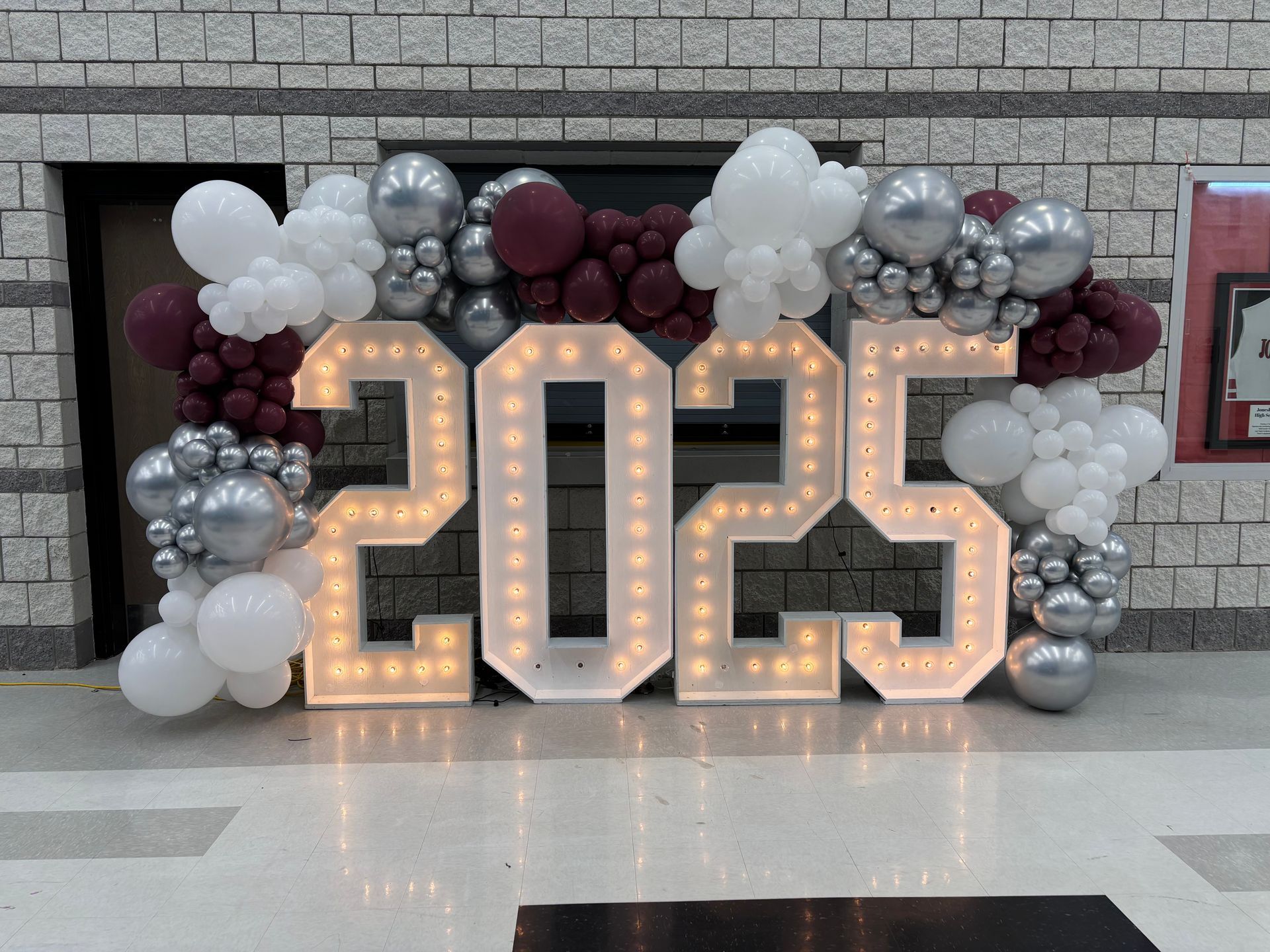 White, silver, and burgundy balloon arch with illuminated 2025 marquee numbers indoors
