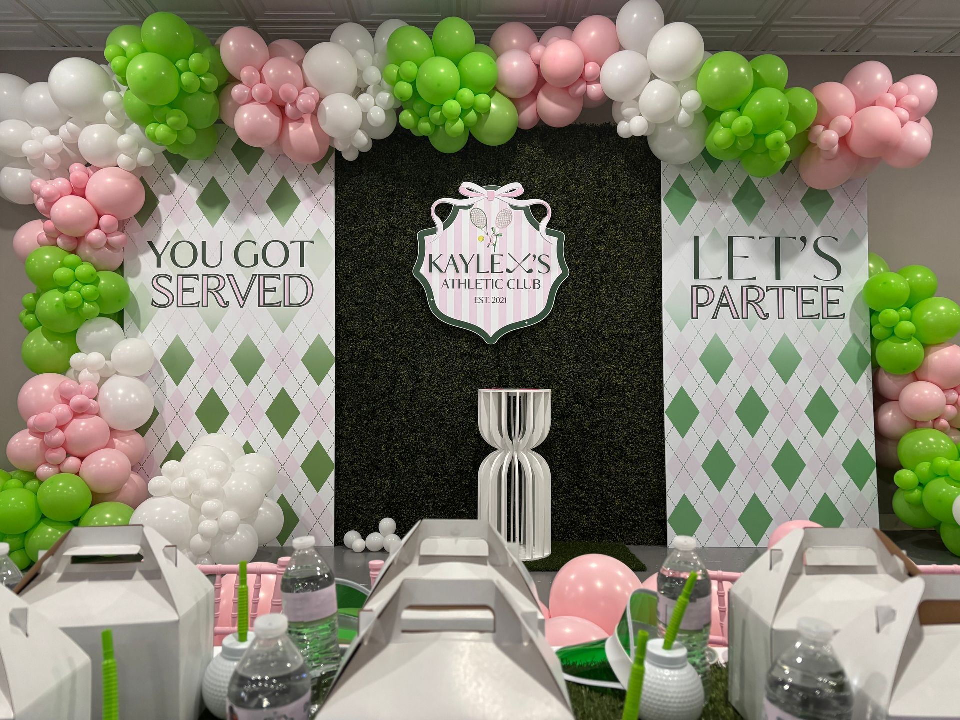 Green hedge wall backdrop with pink, white, and lime balloon garlands and themed party signage for kids birthday photo area