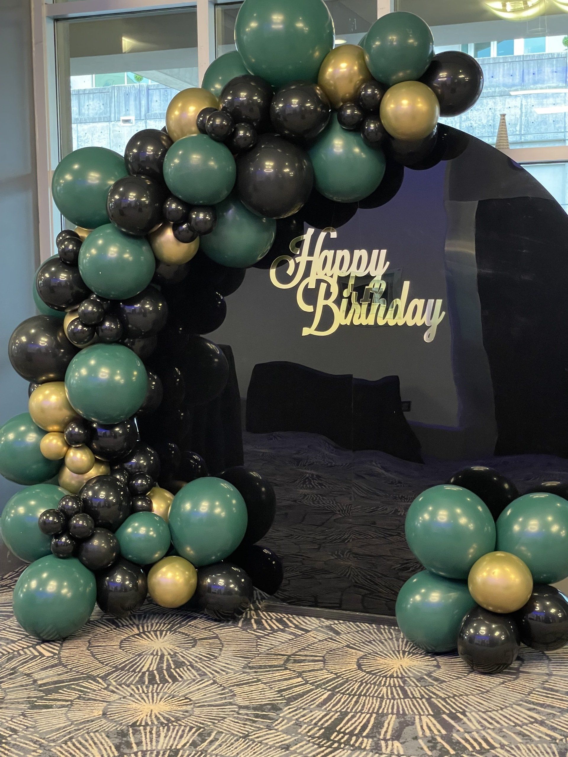 Black teal and gold organic balloon arch with happy birthday sign for modern indoor celebration