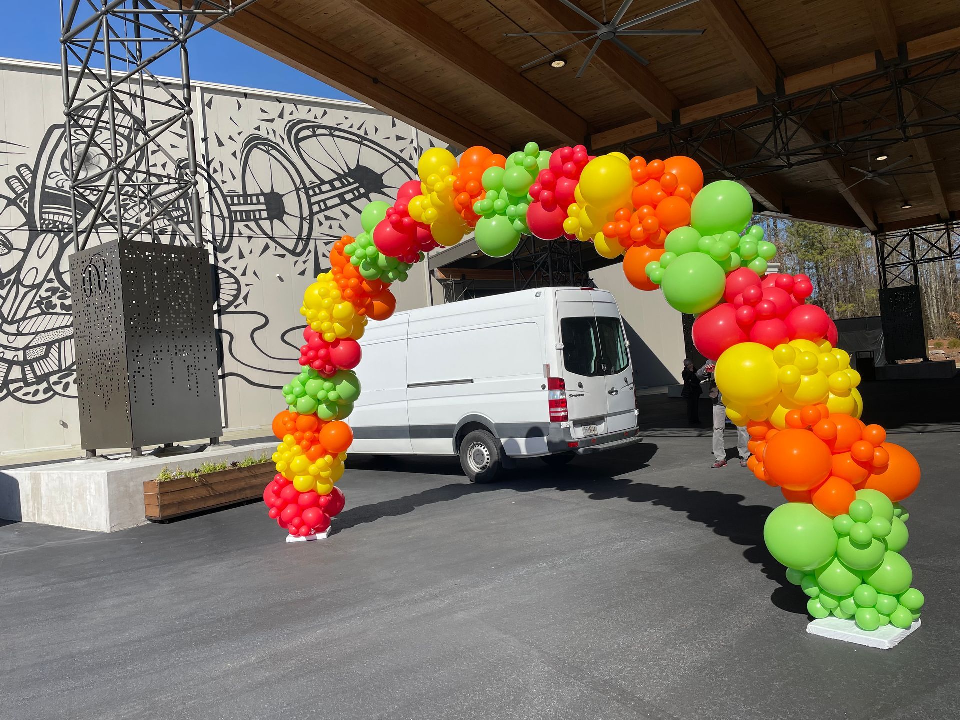 Bright multicolor balloon arch outdoor setup beside a mural and parked van