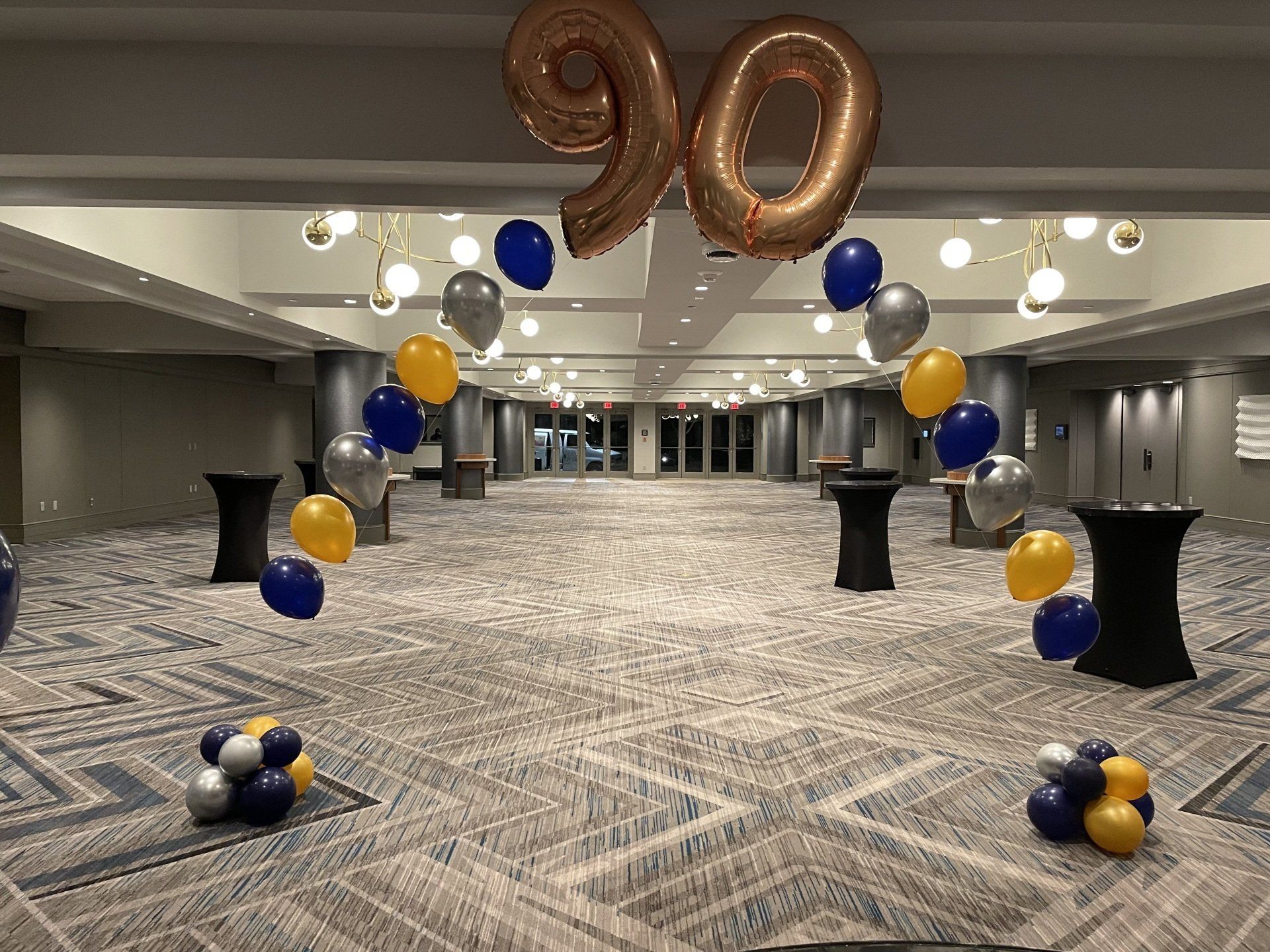 Gold, navy, and silver graduation balloon arch with large number 90 display inside a venue