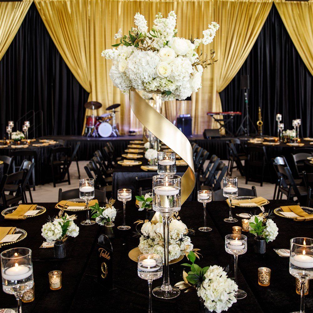Tall white floral centerpiece on a gold stand in front of an elegant stage with gold drapery
