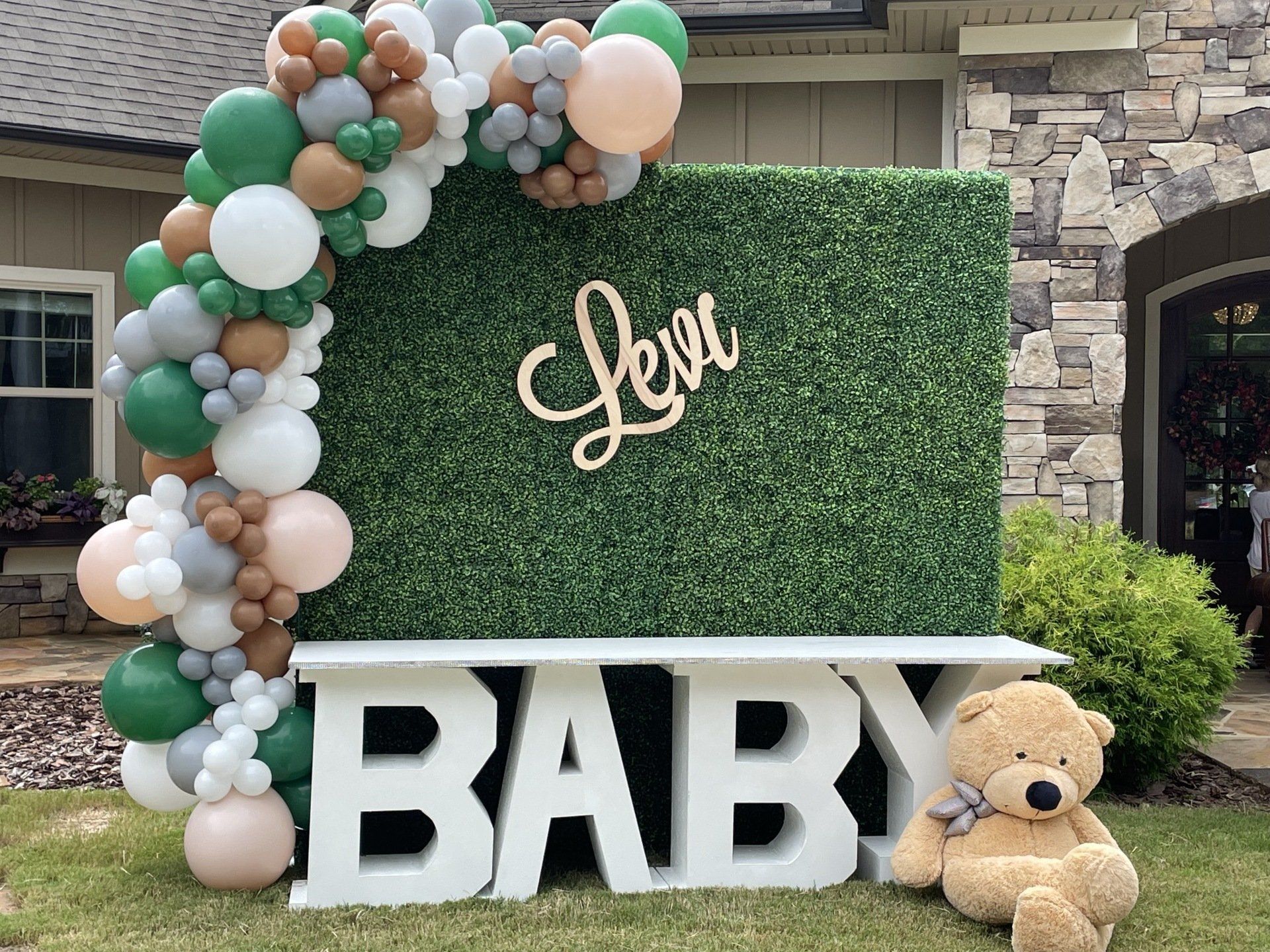 Green white and tan balloon garland framing hedge wall backdrop with custom name sign for baby shower