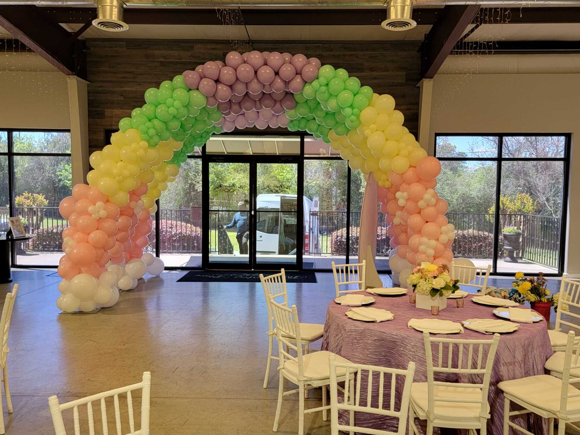 Pastel rainbow balloon arch inside an event space framing a round dining table setup