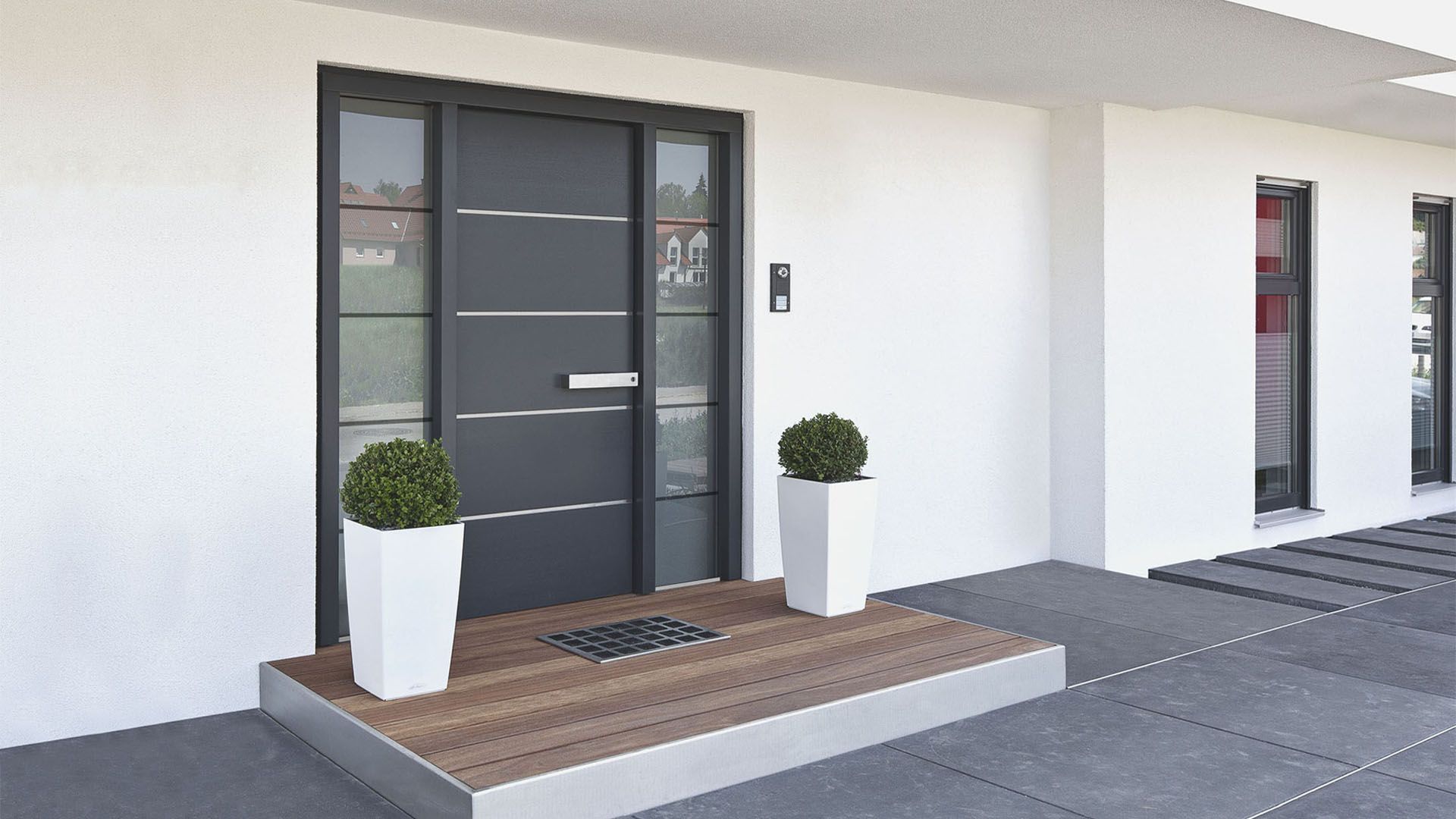 Modern gray front door with side glass panels and two white planters with greenery, set on a wooden platform.
