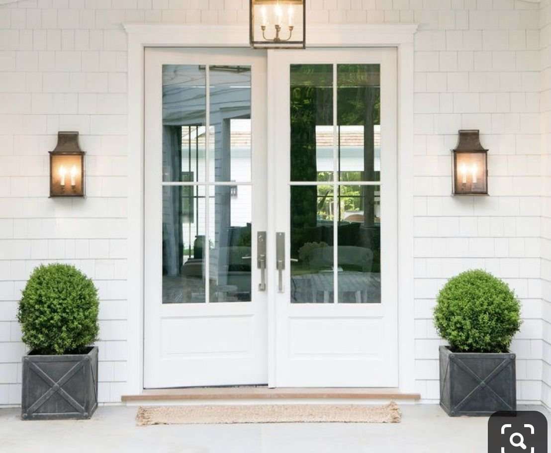 White double doors with glass panes, flanked by lanterns and topiaries in dark square planters.