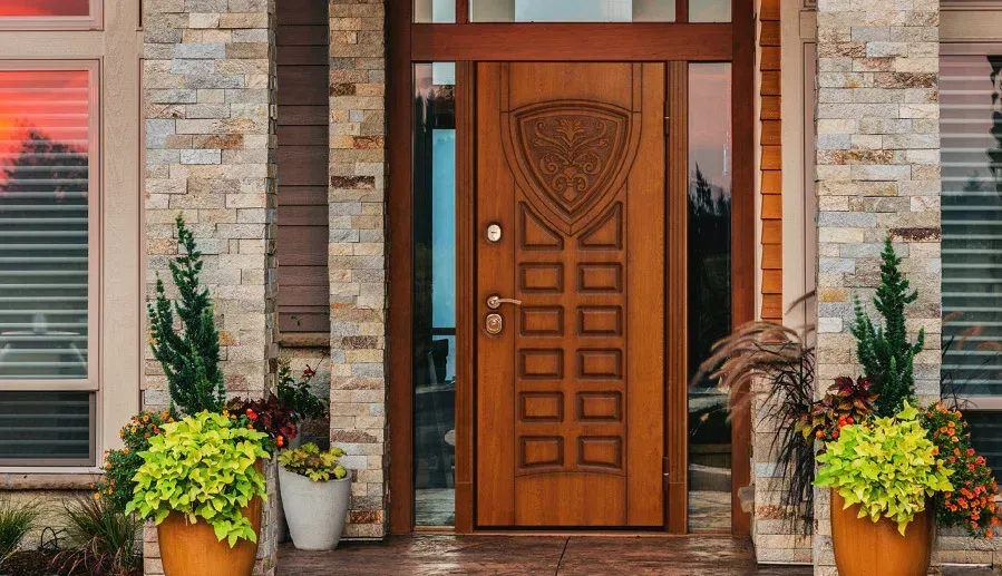 Wooden front door with a decorative shield design, flanked by stone walls and plants in pots.