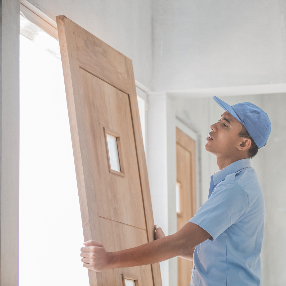 A person in a blue uniform installing a wooden door in a bright white room. He is focused on the task.