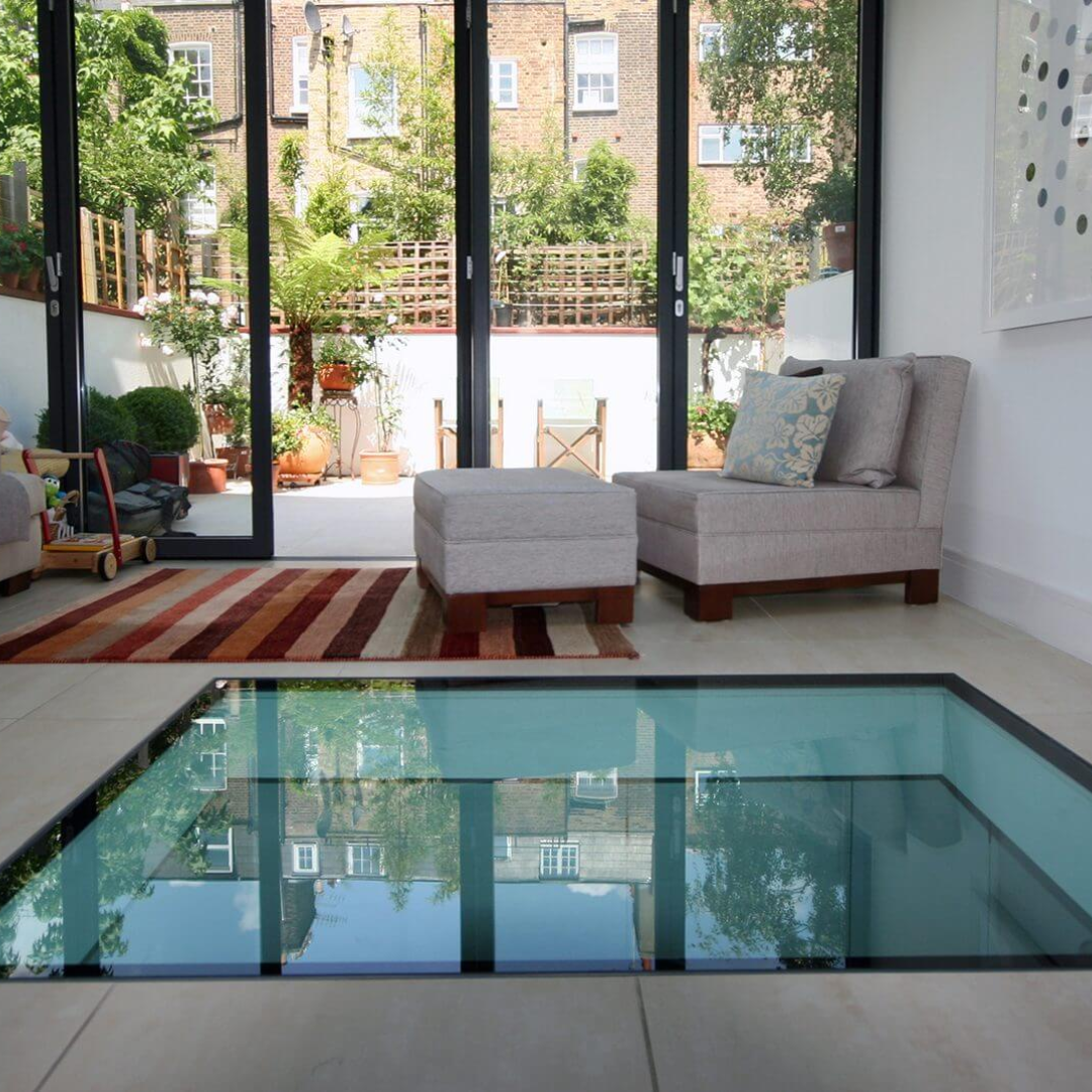 Living room with a glass floor, open to a patio. Neutral furniture sits on a rug, while reflections of buildings are seen through the glass.