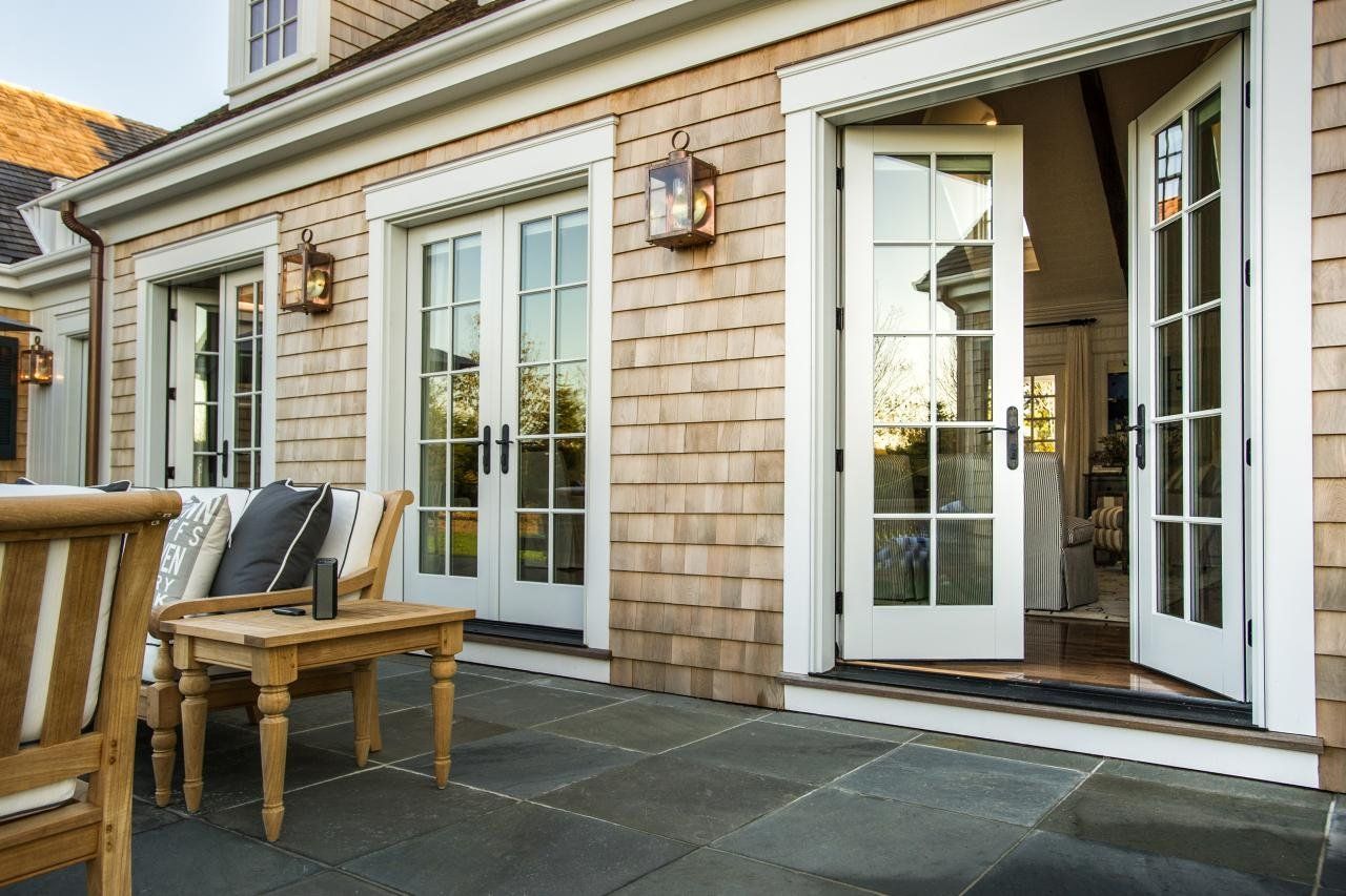 Exterior of a house with weathered wooden shingles, French doors, and a stone patio with outdoor furniture.