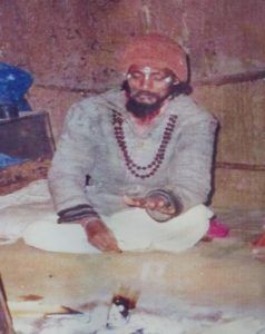A seated person in an orange turban and layered beads, holding a small bowl beside a fire at Enlightened Life Temple