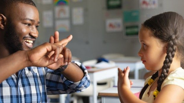 Teacher and child sign to each other in a classroom, smiling across a table.