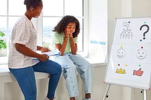 Two people in a bright room discuss a quiz board with icons and a large question mark.