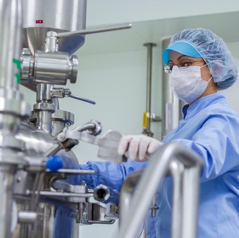 Technician in blue protective gear and mask inspecting stainless steel industrial equipment in a clean lab.