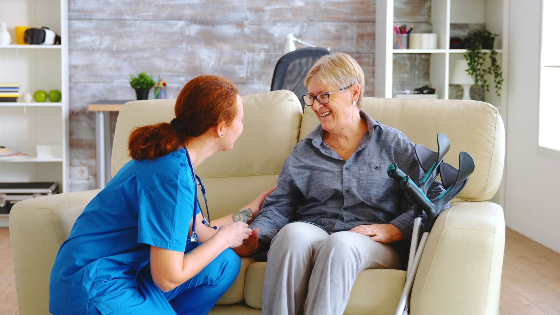 Caregiver in blue scrubs talks with smiling woman on a couch in a bright living room