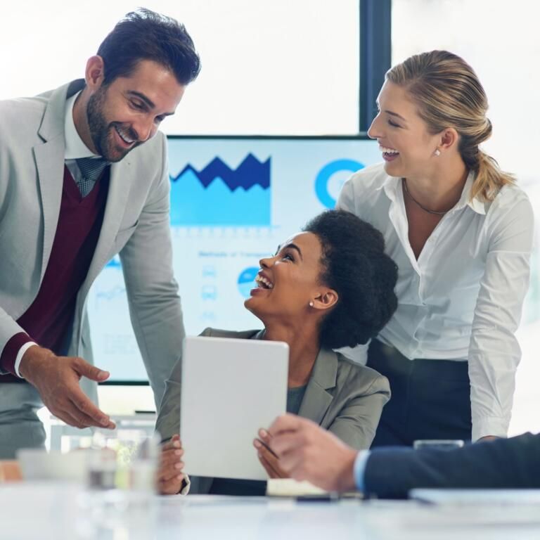 Colleagues smiling and discussing a presentation in a bright office, with charts visible on a screen.
