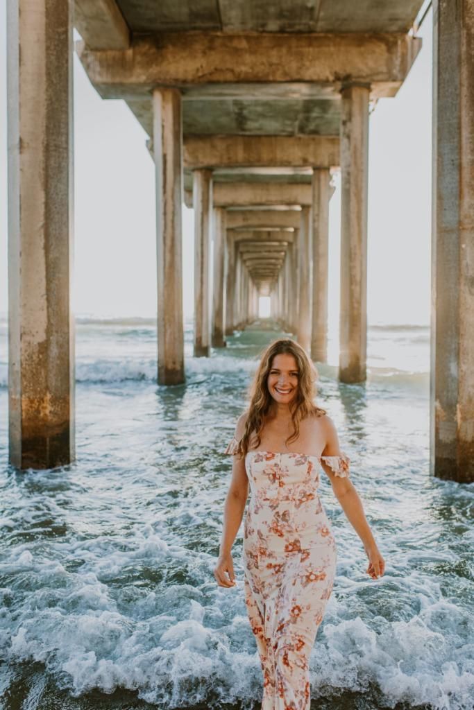 Woman in a floral dress walking through surf under a pier at the beach