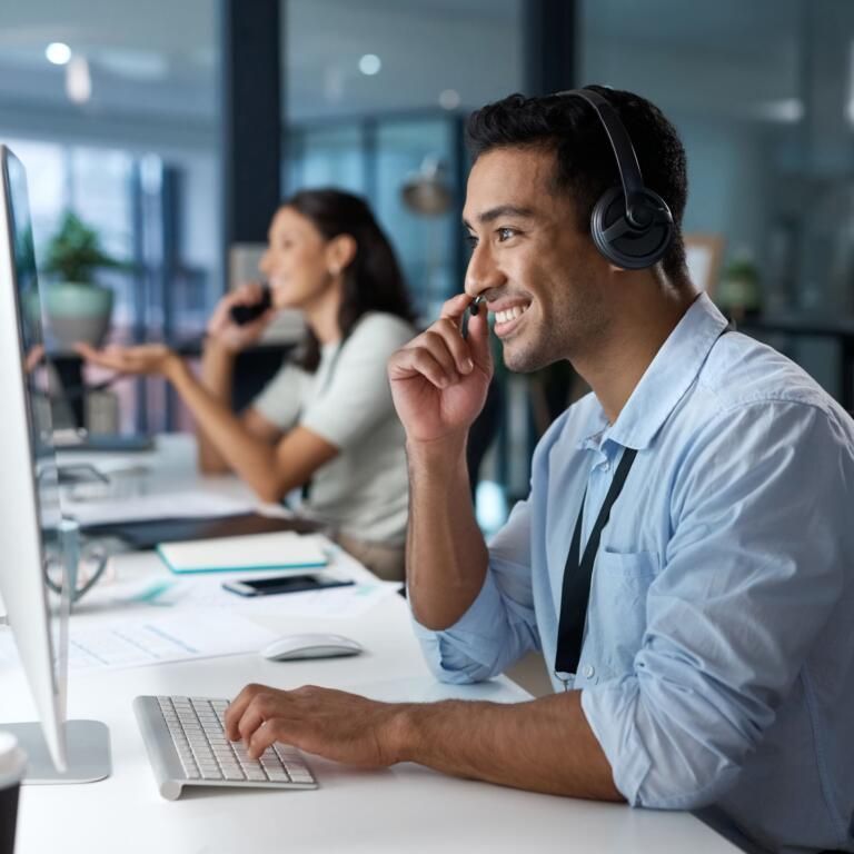 Man in headset smiling at desk in bright office, typing and speaking on phone