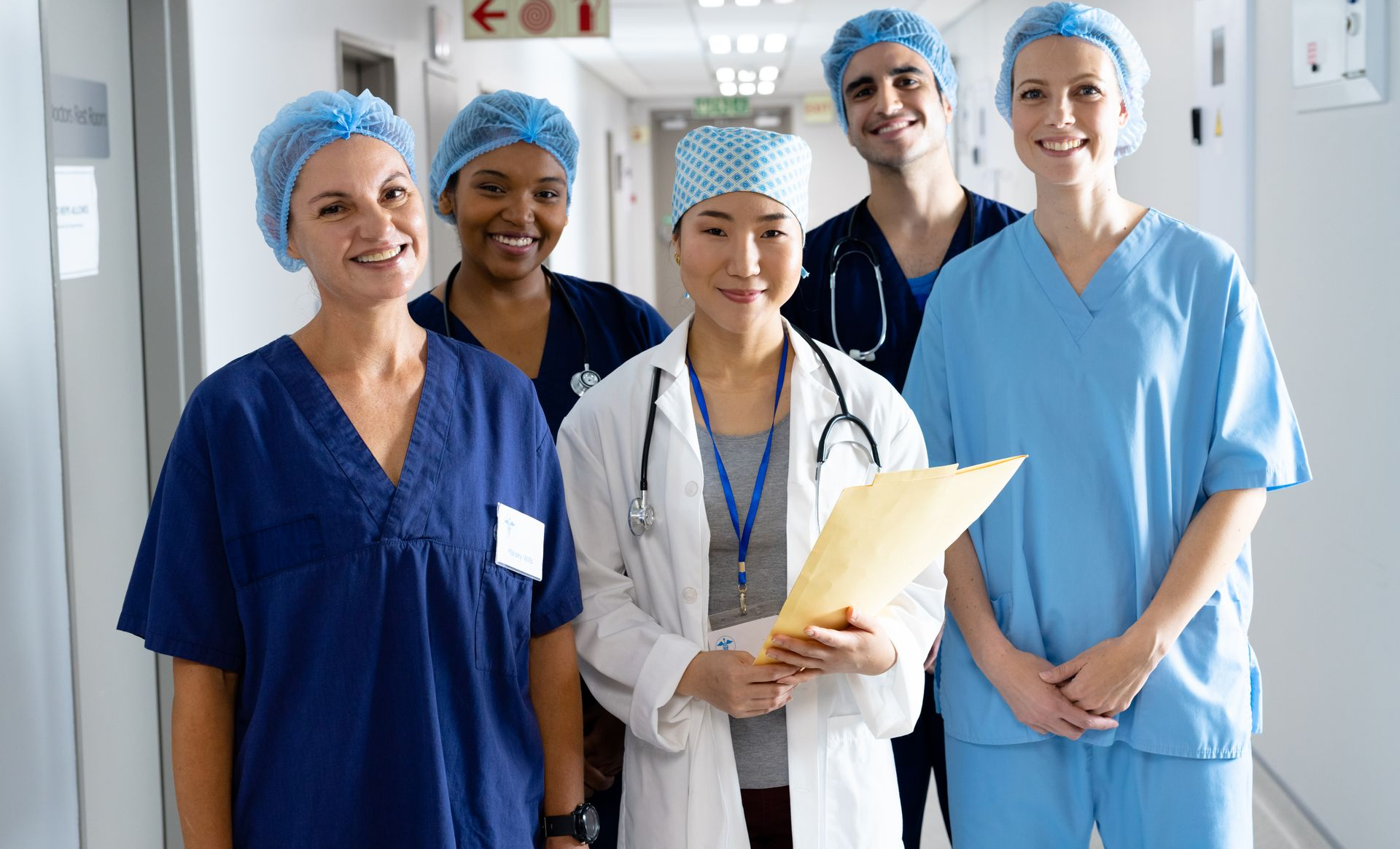Five healthcare workers in scrubs and lab coat standing in a hospital hallway, smiling and holding a chart