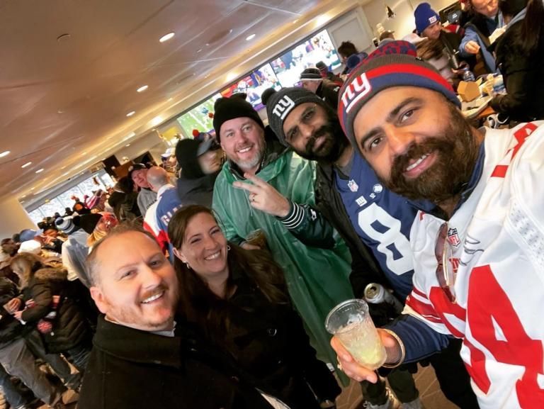 Group of smiling sports fans taking a selfie indoors, wearing jerseys and winter hats, holding drinks.