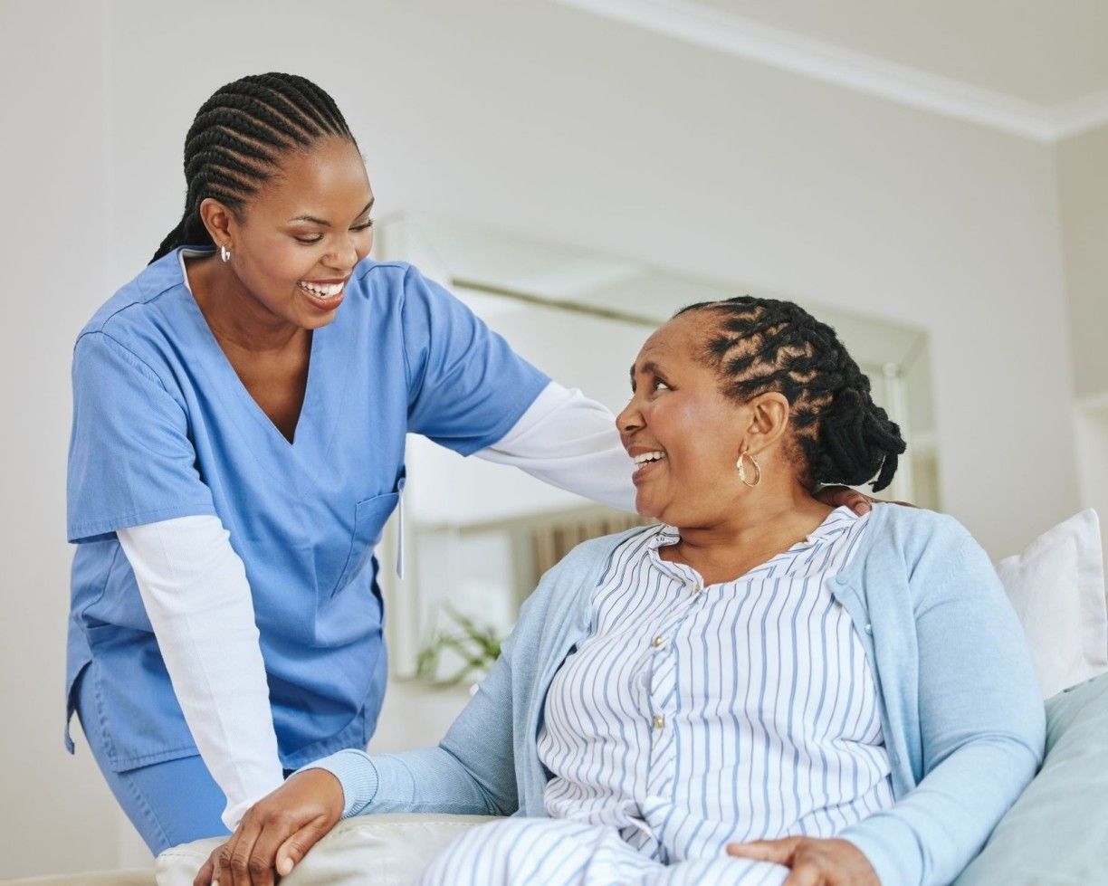 Caregiver in blue scrubs smiling and comforting a seated patient in a bright room