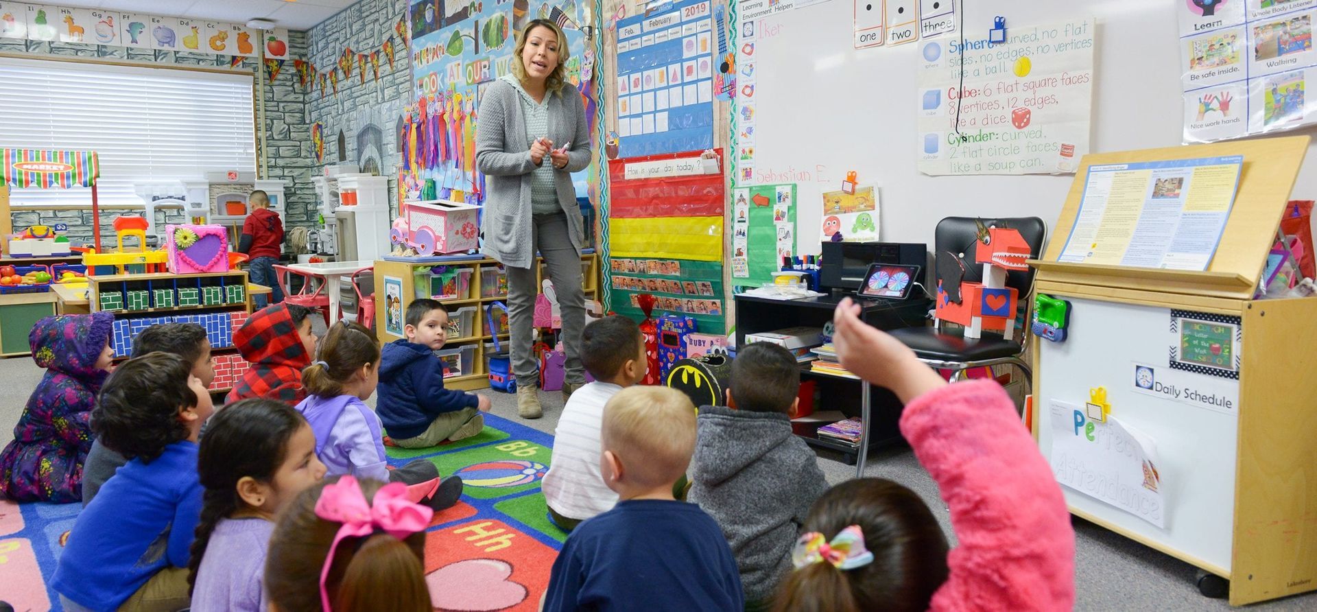 Teacher leading a seated classroom lesson, with children raising hands on colorful rug near whiteboard and bookshelves