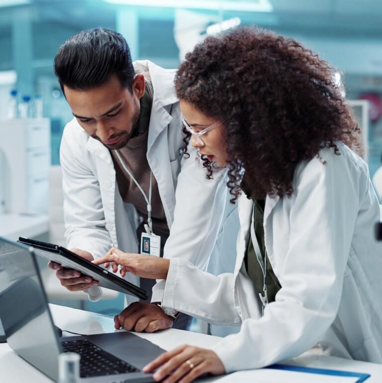Two lab researchers reviewing data on a tablet beside a laptop in a bright lab
