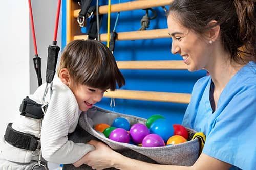 Child on a therapy swing reaching into a tray of colorful balls while a therapist smiles nearby