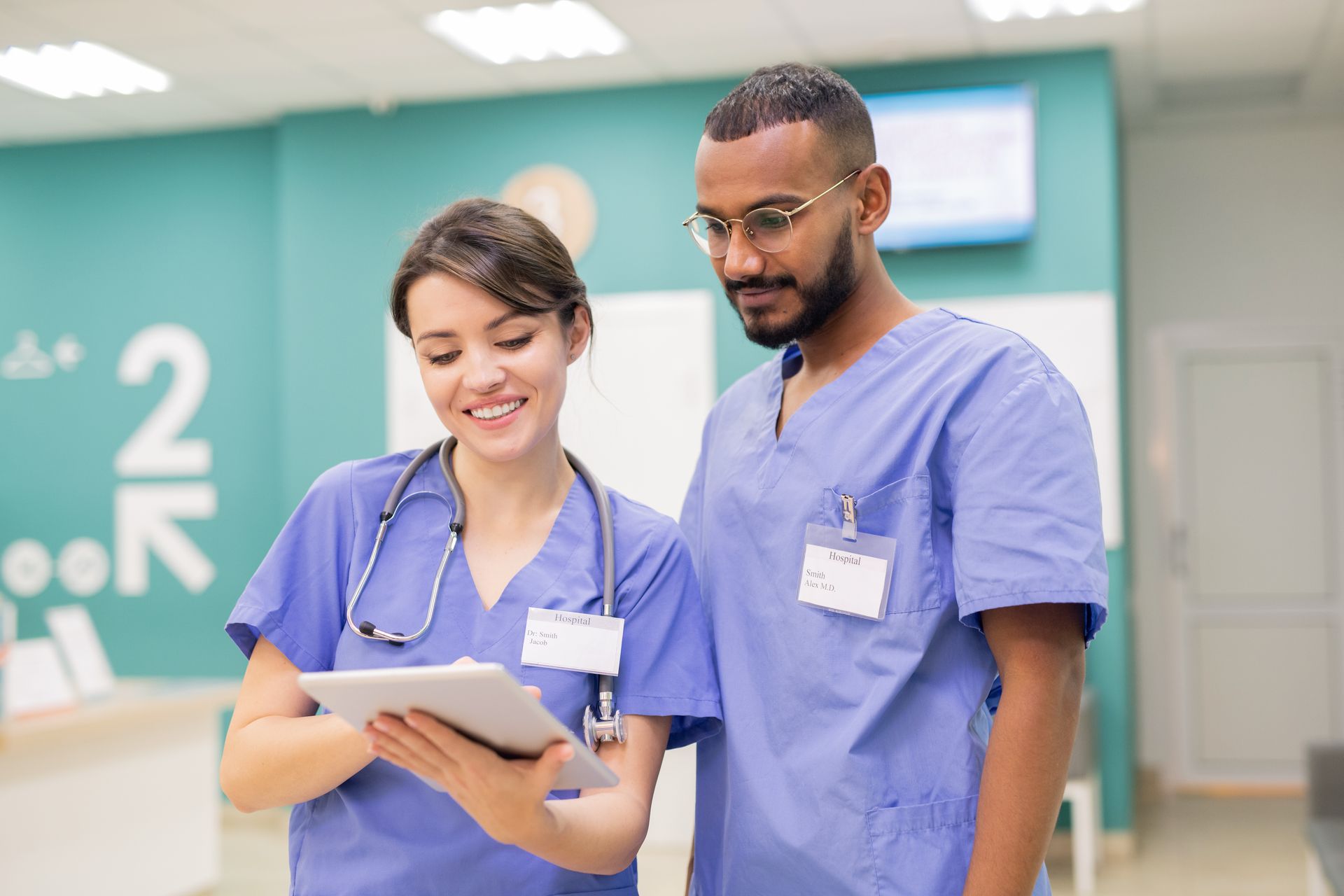 Two healthcare workers in blue scrubs reviewing a clipboard in a hospital hallway