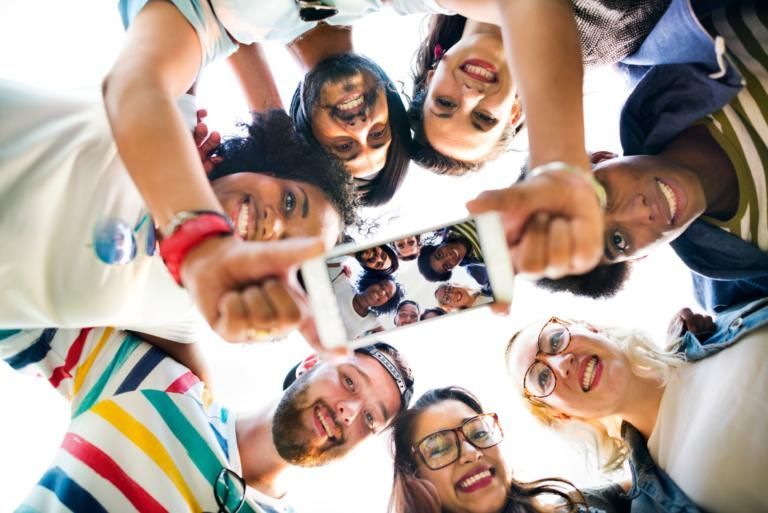 Group of people smiling in a circle, holding a smartphone together outdoors