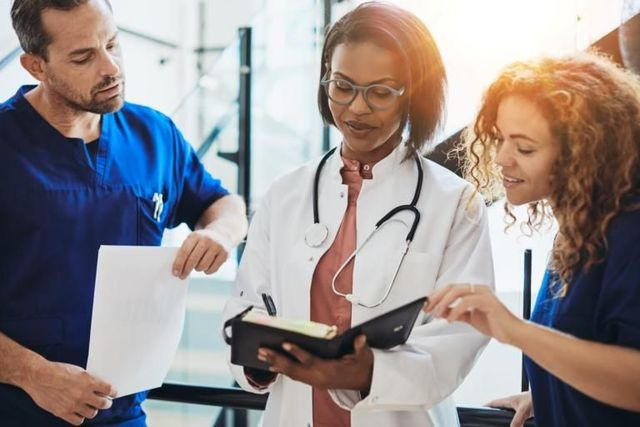 Three clinicians reviewing a tablet and documents in a bright hospital hallway