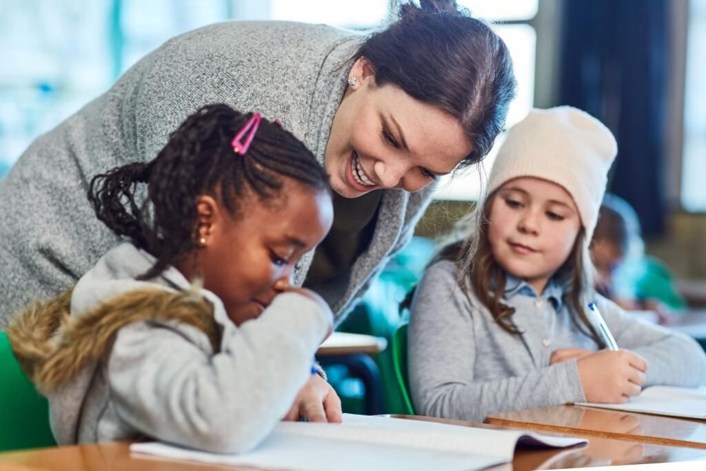 Teacher helping two children write at a table in a classroom