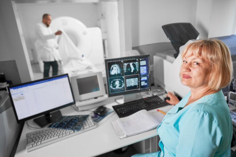 Medical technician at a workstation reviewing scan images while a patient enters the imaging room