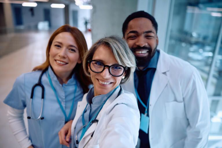 Three smiling medical professionals in white coats and scrubs posing in a hospital hallway