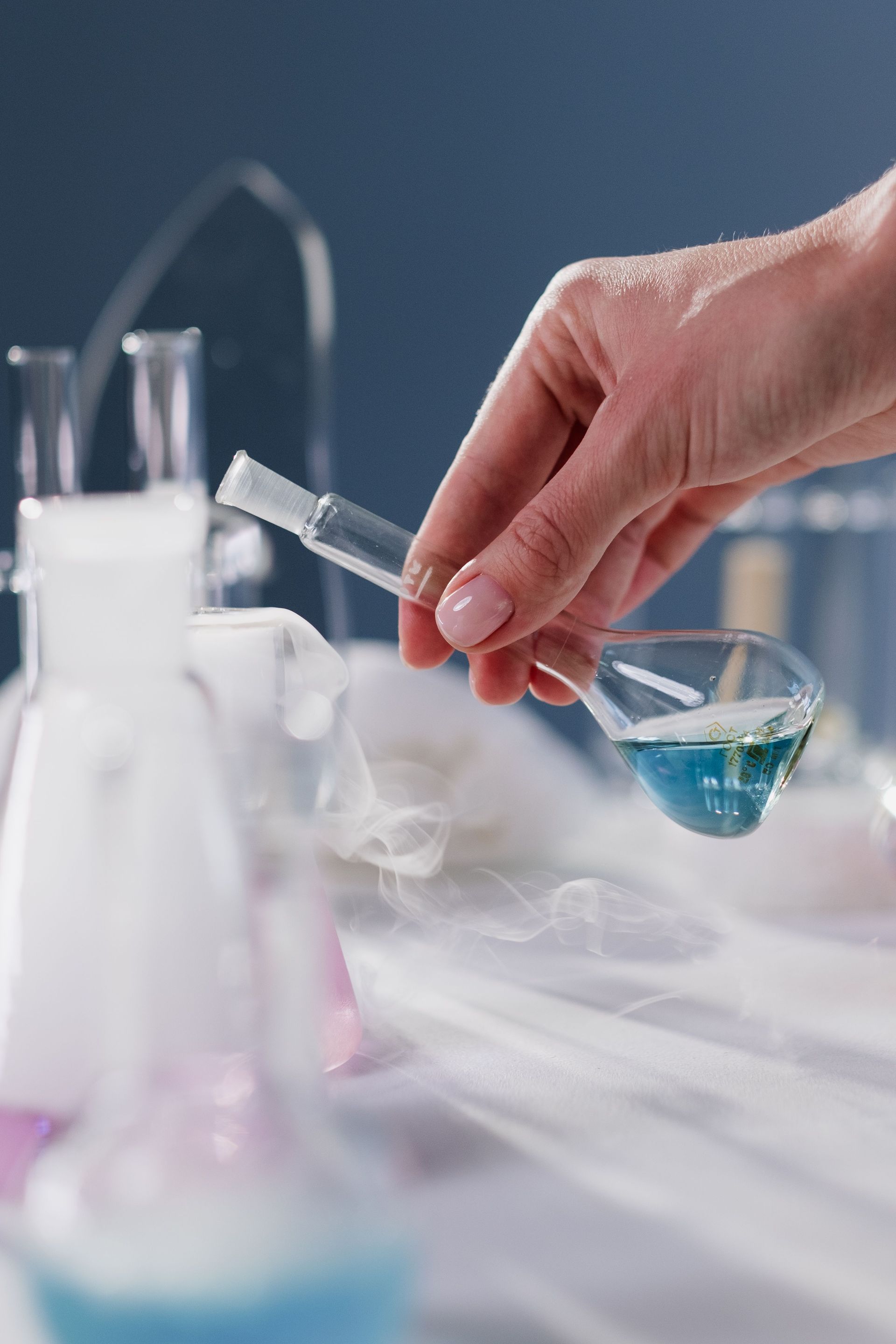 Hand holding a pipette over a beaker in a lab with blue liquid and mist