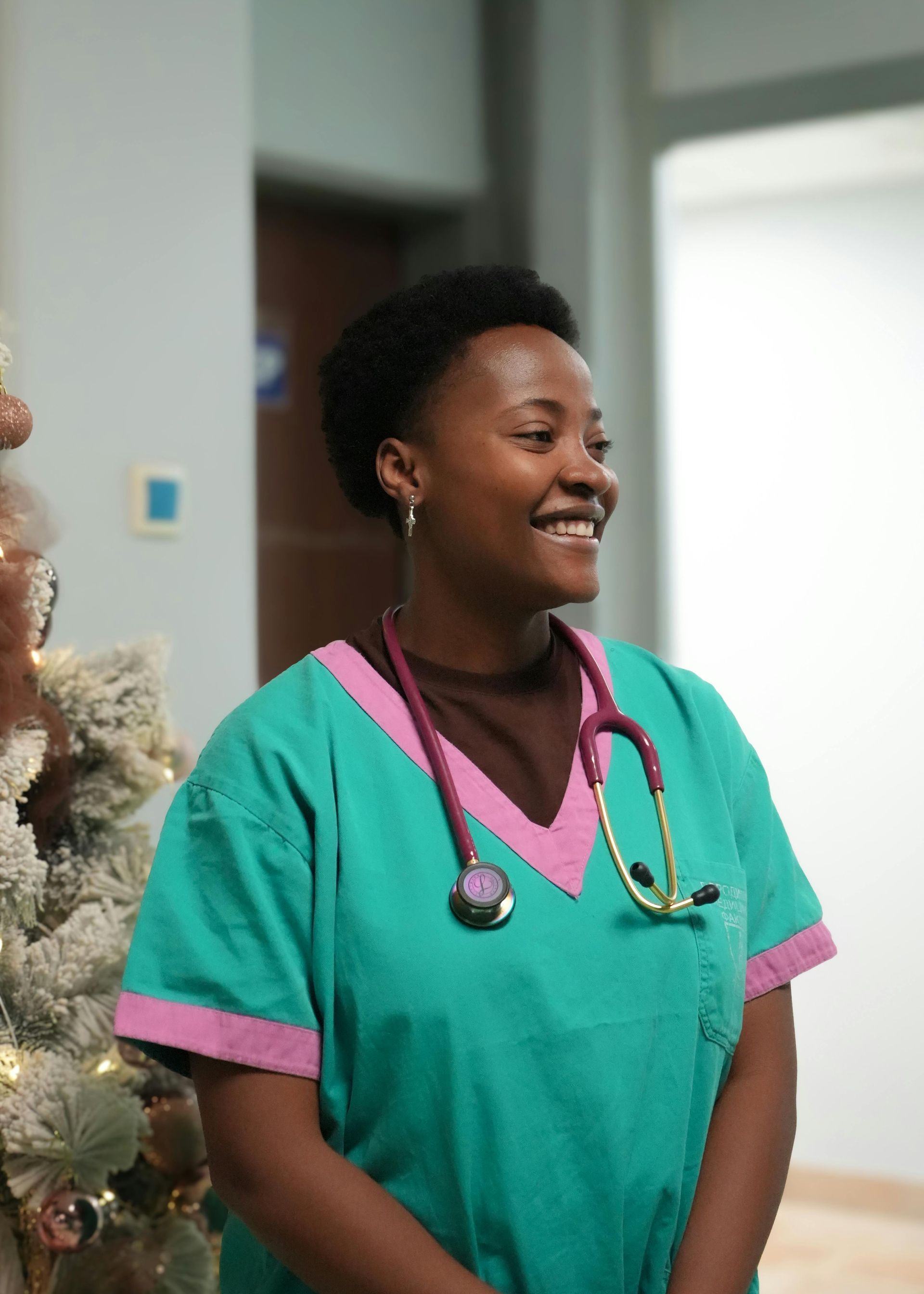 Smiling nurse in teal scrubs with pink trim and stethoscope in a hospital hallway