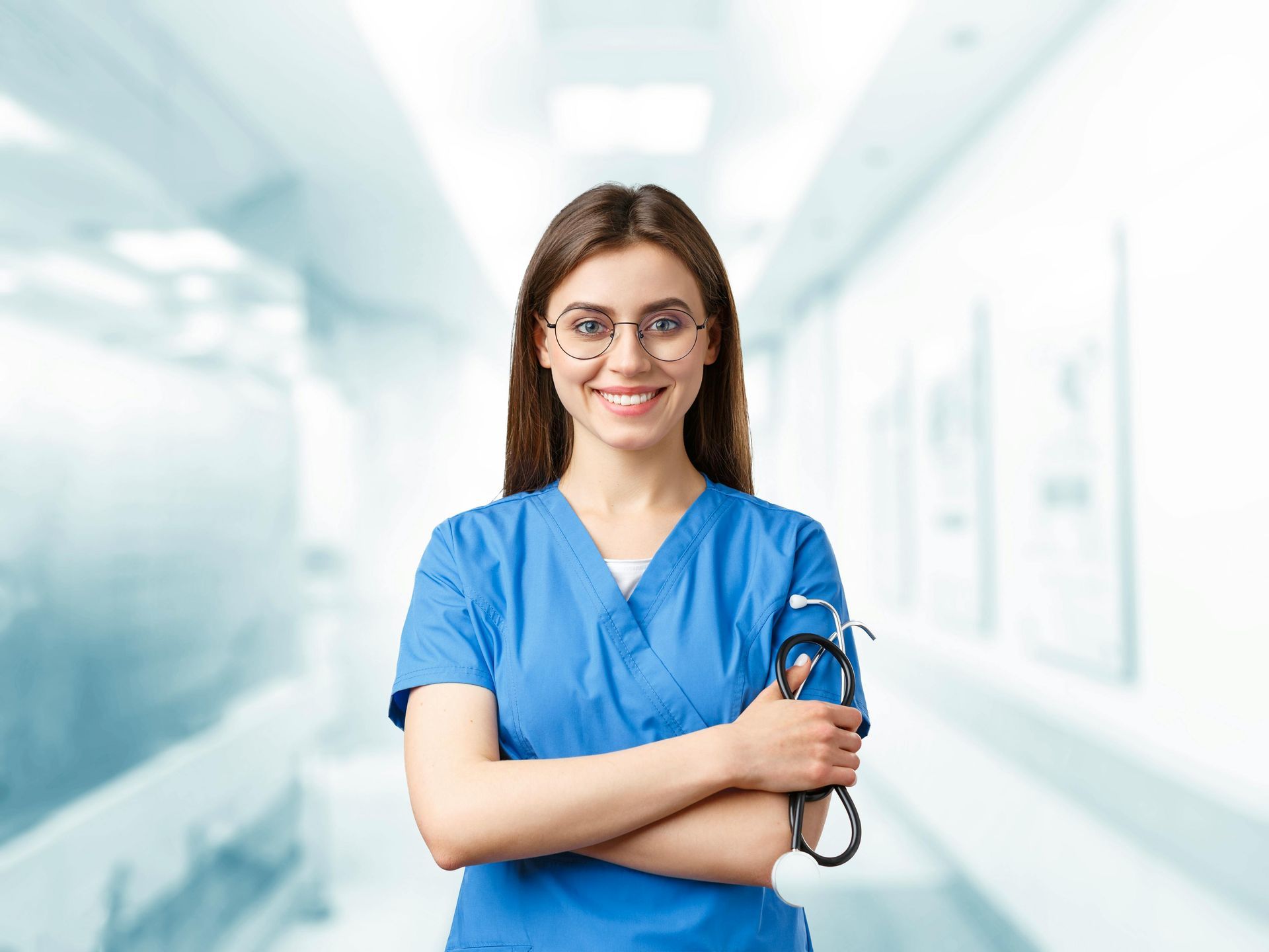 Healthcare worker in blue scrubs holding a stethoscope, standing in a bright hospital hallway.