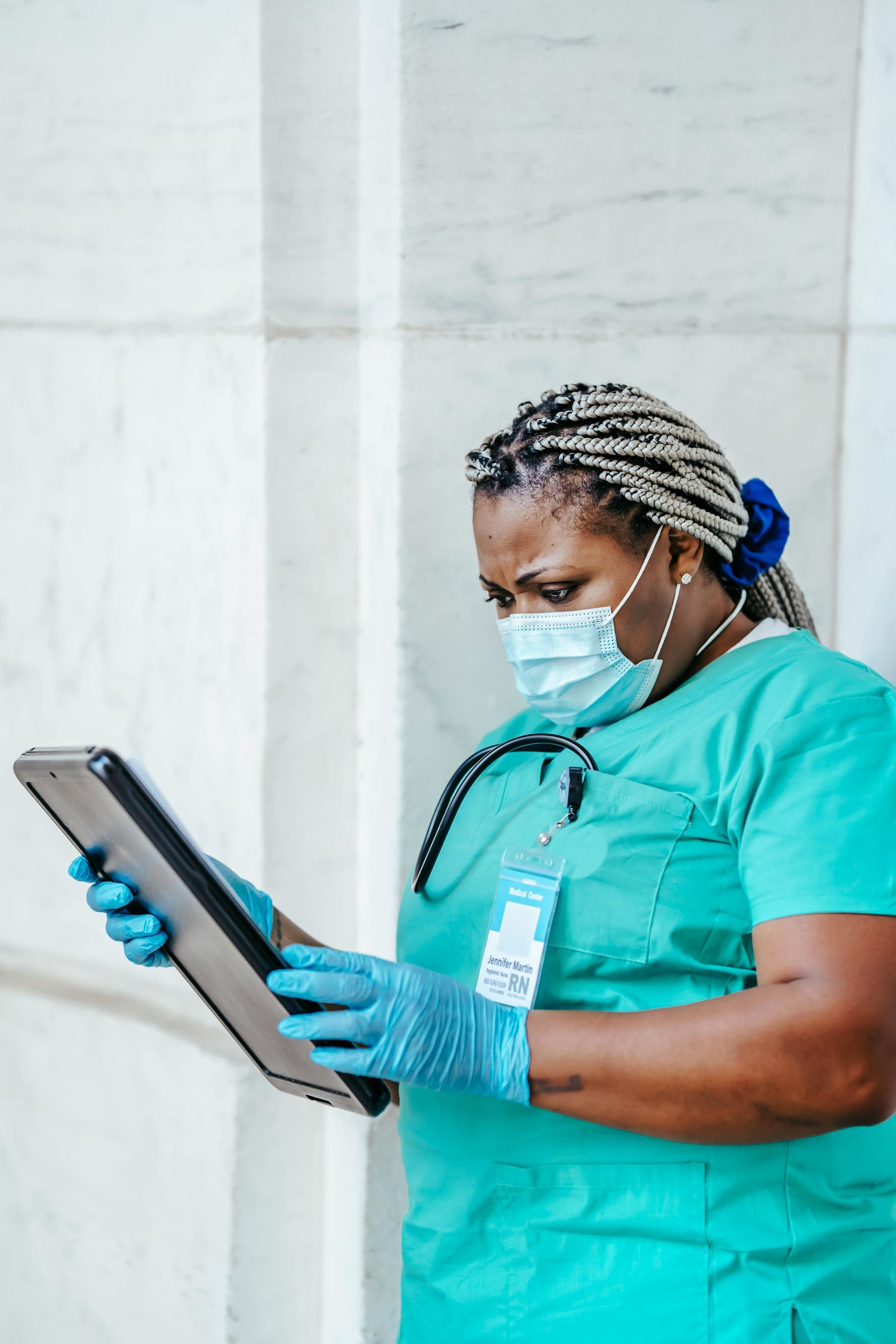 Healthcare worker in teal scrubs, mask, and gloves using a tablet in a clinical setting