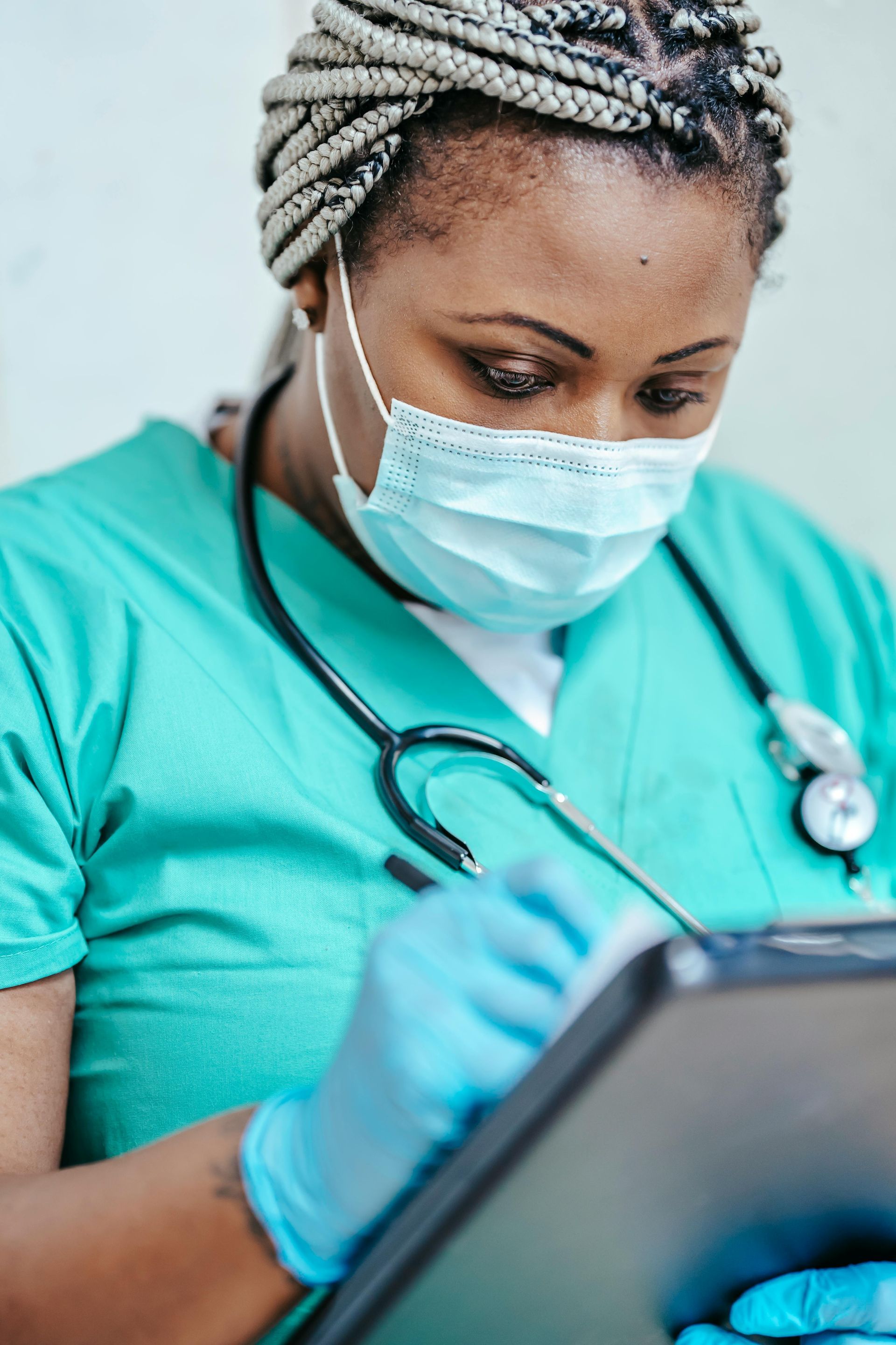 Healthcare worker in teal scrubs and mask writing on a tablet with a stethoscope around her neck