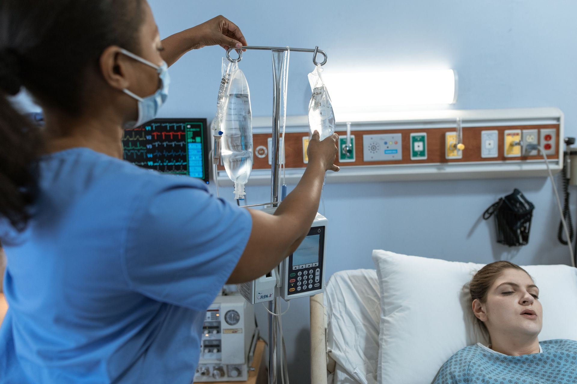 Nurse adjusting IV drip beside a patient resting in a hospital bed with monitors nearby