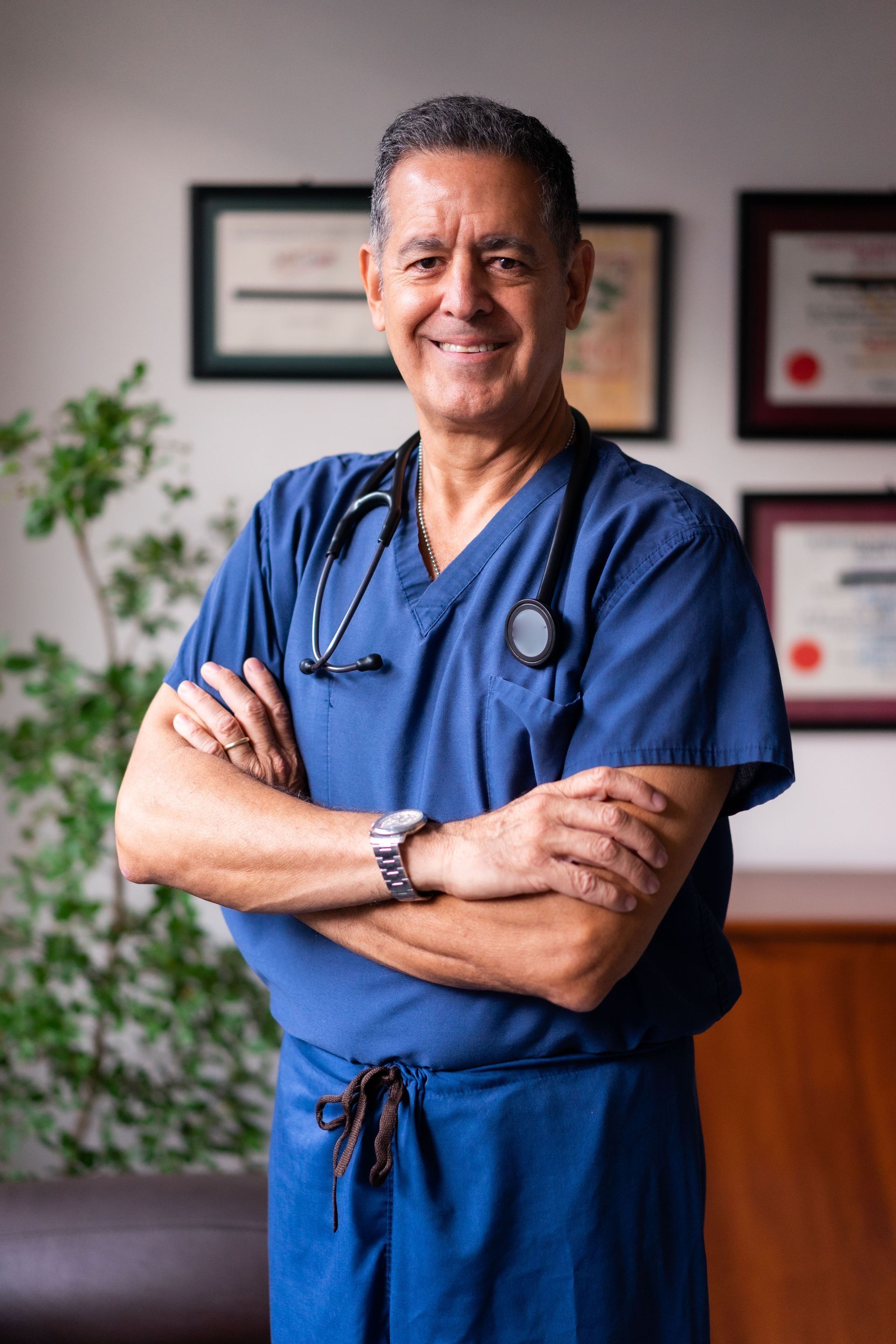 Smiling medical professional in blue scrubs, arms crossed, standing in a clinic office.