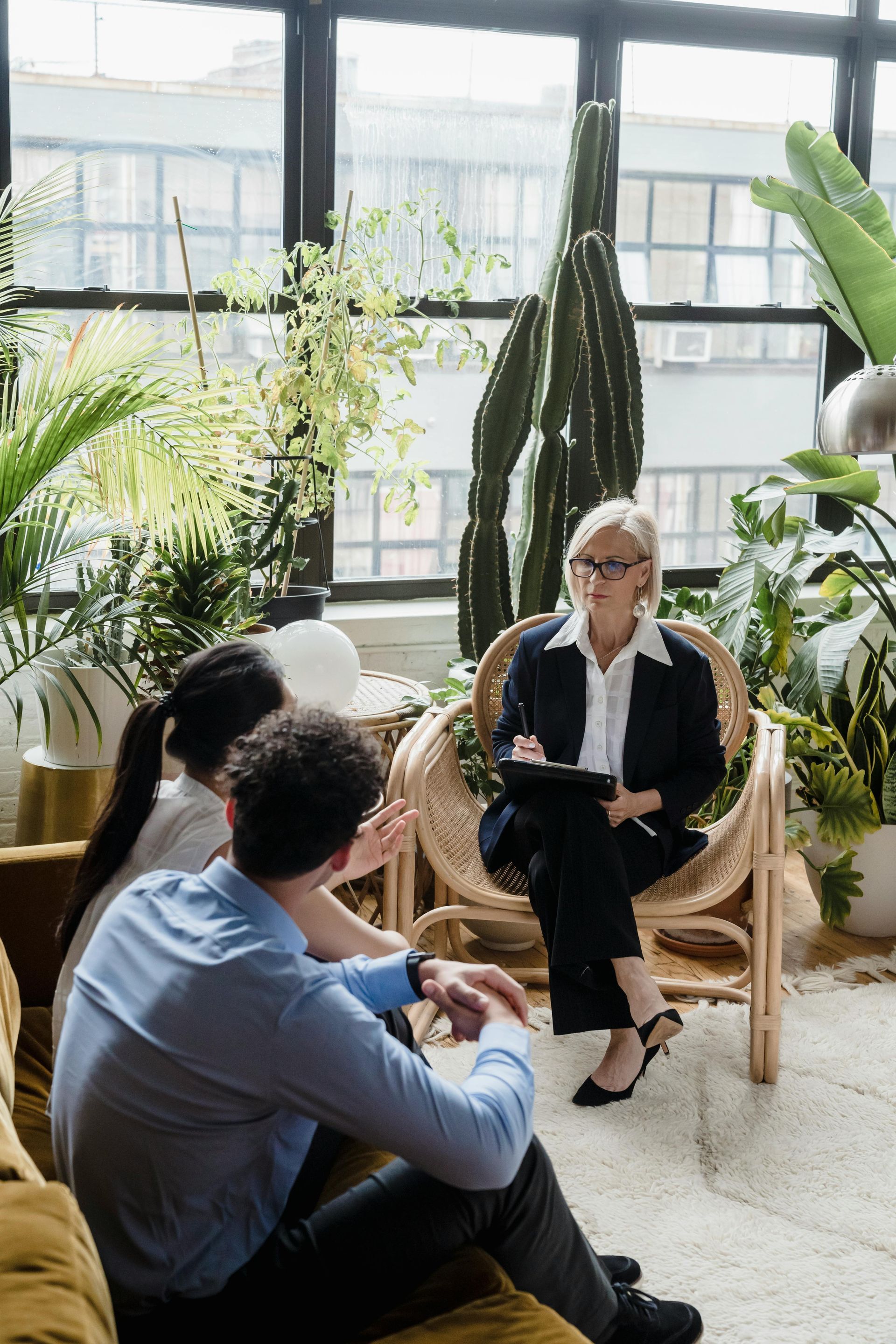 Two people in a bright greenhouse-like room listening to a seated speaker among large plants.